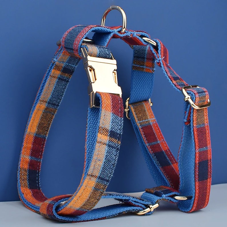May include: A dog harness with a plaid pattern in shades of blue, orange, and burgundy. The harness features gold-colored metal hardware, including a buckle and adjustable rings. The harness is designed for comfort and security.