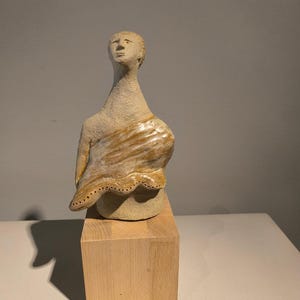 Könnte beinhalten: Eine Keramikskulptur einer menschlichen Büste mit einer texturierten, matten beigen Oberfläche. Die Figur ist mit einem glänzenden, marmorierten braun-weißen stoffartigen Element drapiert, das eine Reihe kleiner, gleichmäßiger Löcher aufweist.