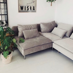 May include: A gray sectional sofa with a light gray throw pillow. The sofa is in a living room with a white wall and a poster with the word "WISTA" on it. There are two hanging plants in the background.