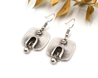 May include: A pair of silver-toned metal drop earrings featuring a square-shaped plate with a central U-shaped cutout. A small metal sphere hangs from the center of each cutout, suspended by a thin link, attached to a standard silver-toned ear wire.