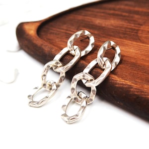 Pode incluir: Um par de brincos pendentes com um design de corrente vertical composto por três elos ovais entrelaçados. Cada elo é feito de metal prateado com um acabamento martelado texturizado que cria uma aparência facetada em toda a superfície.