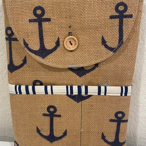 May include: A burlap tote bag with a flap closure, featuring a navy blue anchor pattern. The bag has a white and navy striped accent and a wooden button closure. The overall design has a nautical theme.