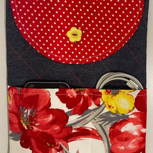 May include: A quilted denim tablet sleeve with a red polka dot flap secured by a yellow flower-shaped button. The front pocket features a floral print with red, yellow, and gray flowers. A phone and charging cable are visible in the pockets.
