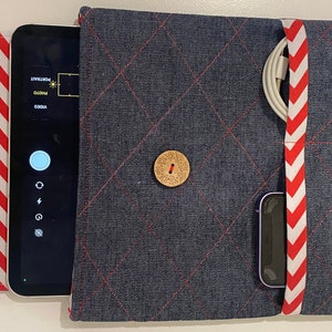 May include: A denim tablet case with red stitching and a wooden button closure. The case holds a tablet, a charging cable, and a smartwatch. A red and white chevron patterned flap is on the left side. The tablet screen displays the camera interface.