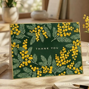Peut inclure: Carte de remerciement avec un fond vert foncé et une bordure dorée. Elle présente un motif floral de fleurs de mimosa jaunes et l'inscription "THANK YOU" en blanc. Un petit vase de fleurs de mimosa est à proximité.