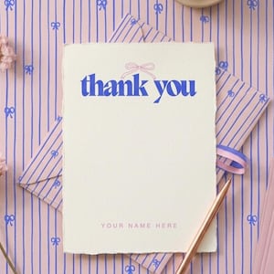 Peut inclure: Une carte crème avec les mots "thank you" en bleu, surmontée d'un nœud rose. La carte est sur un fond rose avec des rayures bleues et des motifs de nœuds. Un stylo doré et des enveloppes roses sont également présents.
