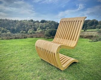 Op de afbeelding: Een houten stoel met een latten rugleuning en zitting. De stoel is gemaakt van natuurlijk hout en heeft een gebogen ontwerp. De stoel staat op een grasveld met een bos op de achtergrond.