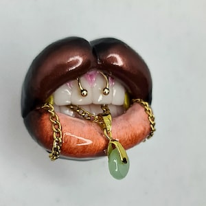 May include: A sculpture of dark brown lips with a glossy finish, featuring white teeth, a gold septum ring, and a gold chain necklace draped over the lower lip. A small, oval-shaped light green pendant hangs from the center of the gold chain.