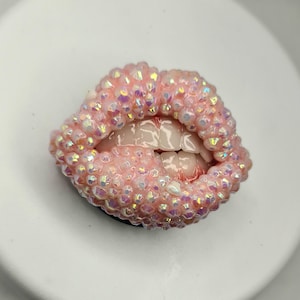 May include: A phone grip accessory shaped like a pair of lips, fully encrusted with iridescent light pink crystals. The center of the lips reveals a set of realistic, off-white teeth, creating a textured, three-dimensional appearance.