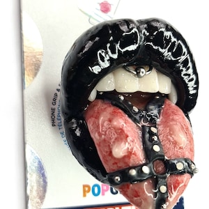 Puede incluir: Labios negros con una lengua que sobresale y una correa de cuero con tachuelas alrededor de la lengua. Los labios tienen un piercing de anillo plateado. El fondo es un empaque de PopSockets blanco y plateado con las palabras "PopSockets" y "Marvel" impresas en él.