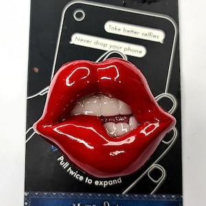 May include: Red glossy lips phone grip with a white tooth showing. The text on the packaging reads "Take better selfies" and "Never drop your phone". The text on the bottom of the grip reads "Pull twice to expand".