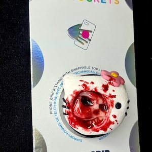 May include: A white phone grip with a bloody skull design and a pink flower on top. The grip is a PopGrip brand and is interchangeable. The text "PHONE GRIP & STAND WITH SWAPPABLE TOP. INTERCHANGEABLE" is printed on the packaging.