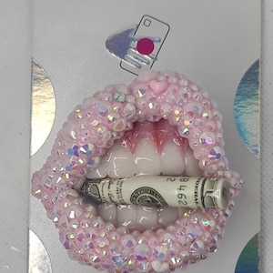 Puede incluir: Un PopGrip PopSockets con un diseño de boca rosa enjoyada, que sostiene un billete enrollado. El PopGrip está sobre un respaldo blanco y plateado, con los logotipos de PopSockets y PopGrip.