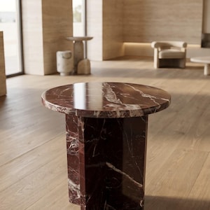 Pode incluir: Uma mesa lateral redonda com um tampo de mármore vermelho escuro e uma base a condizer. A mesa tem uma superfície polida com veios brancos. A base tem uma forma geométrica. A mesa está sobre um chão de madeira clara.