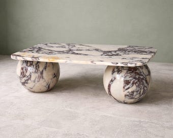 May include: A rectangular coffee table with a marble top and two spherical bases. The marble has a cream background with dark purple veining. The table is a unique piece of furniture.