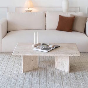 Puede incluir: Una escena de salón de tonos neutros con un sofá blanco con cojines marrones y beige. Una mesa de centro rectangular de piedra sostiene una revista abierta y dos velas blancas. Una alfombra color crema y una lámpara pequeña completan la decoración.