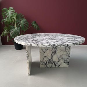 Op de afbeelding: Een ronde marmeren salontafel met een wit en grijs geaderd patroon. De tafel heeft een vierkante basis en een enkele rechthoekige steun. Een plant in een pot staat op de achtergrond.