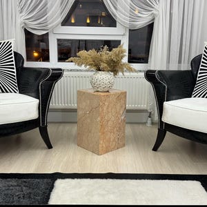 May include: A living room scene featuring two black armchairs with white cushions and decorative pillows. A square marble side table holds a decorative vase with dried flowers. The room has sheer white curtains and a patterned rug.