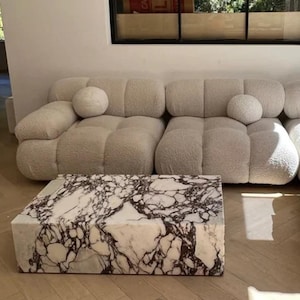 May include: A contemporary living room arrangement with a cream-coloured, modular sofa with rounded cushions and two matching round throw pillows. A rectangular coffee table with a white marble top and dark brown veining sits in front.