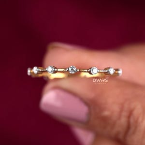 Könnte beinhalten: Ein zarter Goldring mit einer Reihe kleiner, klarer Diamanten, die in einem schlichten Design gefasst sind. Der Ring wird an einem Finger gezeigt.