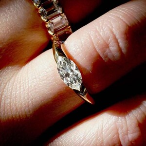 Puede incluir: Un anillo de oro con un diamante talla marquesa. El diamante es el punto focal, engastado en una banda moderna y elegante. El anillo se sostiene entre los dedos, mostrando su brillo y dise&ntilde;o. El anillo es parte de una colecci&oacute;n de joyas.