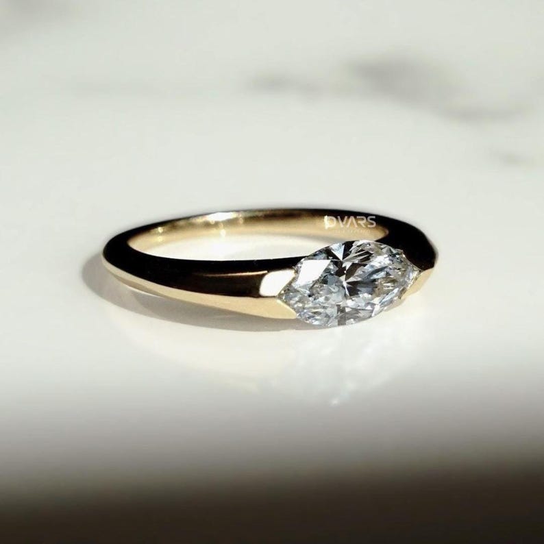 Puede incluir: Un anillo de oro con un diamante talla marquesa. El diamante est&aacute; engastado en un bisel y la banda es lisa y pulida. El anillo est&aacute; sobre una superficie blanca. La palabra "DVARS" est&aacute; grabada en la banda.