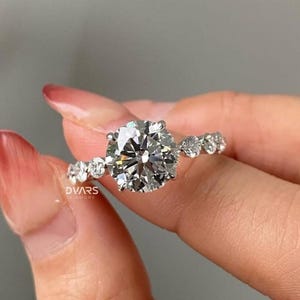 Puede incluir: Un anillo de diamantes brillante con una piedra central grande y redonda y piedras más pequeñas en la banda. El anillo es sostenido por una mano, mostrando su brillo. El anillo es una joya.