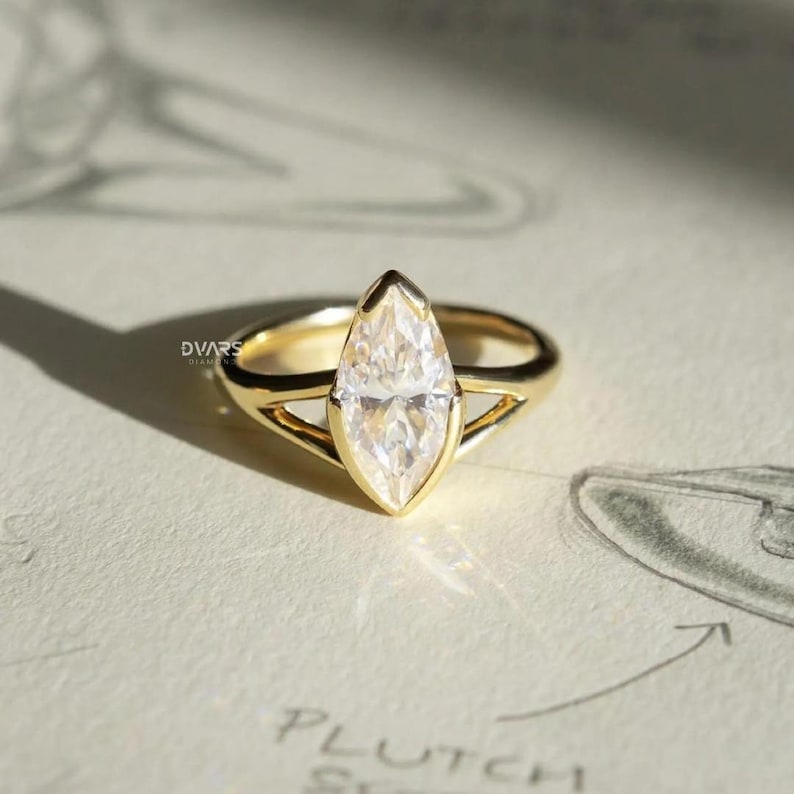Pu&ograve; includere: Un anello d'oro con un diamante taglio marquise. Il diamante &egrave; incastonato in una montatura a castone e la fascia ha un design a gambo diviso. L'anello &egrave; su una superficie bianca con uno schizzo sullo sfondo. L'anello &egrave; di Dvars Diamonds.