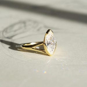 Pu&ograve; includere: Un anello d'oro con un diamante taglio marquise. Il diamante &egrave; incastonato in una montatura a castone e la fascia ha un design a gambo diviso. L'anello &egrave; presentato su una superficie chiara, con il marchio "DVARS" visibile sulla fascia.