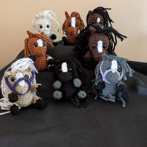 Puede incluir: Una colección de siete figuras de caballos de ganchillo en varios colores. Los caballos tienen crines y colas, algunos con detalles morados o blancos. Los caballos están sobre una superficie negra.