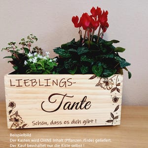 Könnte beinhalten: Ein Holz-Pflanztopf mit dem Text "LIEBLINGS-Tante Schön, dass es dich gibt!" Der Topf ist mit Blumenmustern verziert und enthält eine rote und grüne Pflanze.