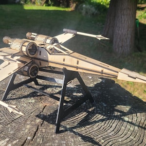 May include: A wooden starfighter model, showcasing intricate wing and engine details. The model is presented on a black stand, with a natural outdoor background. The starfighter is constructed from light-coloured wood, with a detailed design.