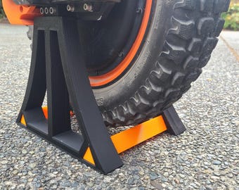 May include: A black and orange stand for a single wheel, designed to hold a unicycle or electric unicycle upright.