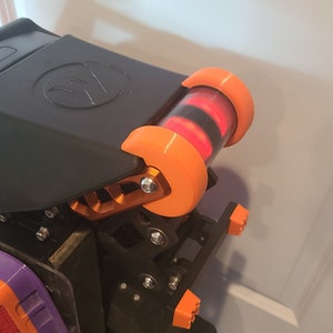 May include: A black and orange 3D printed device with a red light inside a clear tube. The device has a black cover with a logo on it. The device is mounted on a black base with orange and purple accents.
