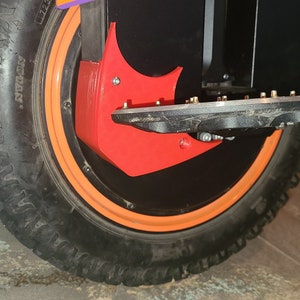 May include: Close-up of an electric unicycle wheel. The tire is black with a deep tread pattern. The wheel has an orange rim and a red protective cover. A black footrest with metal studs is visible.