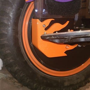 May include: A close-up of a black tire with an orange rim and a black and purple fender. The fender has a flame design and is attached to the tire with screws.