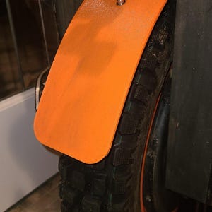 May include: Orange fender with two silver screws attached to a black tire with a tread pattern. The tire is mounted on a black wheel with a red rim.