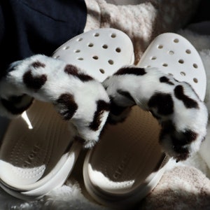 May include: White clogs with a leopard print fur trim. The clogs have a textured sole and the brand name "Crocs" is visible on the bottom.