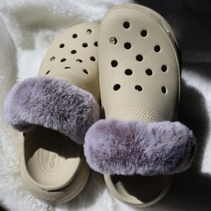 May include: A pair of beige clogs with a purple faux fur lining. The clogs have a classic design with a rounded toe and a comfortable fit.