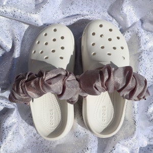 May include: White clogs with a pink satin bow tied around the straps. The clogs have a textured sole and the words "Iconic Comfort Crocs" are printed on the inside.