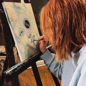 May include: A rectangular canvas painting on a black wooden easel features a stylized face with a prominent black button replacing one eye. The artwork uses shades of pale blue, cream, and white, with a fine brush adding detail to the lips.