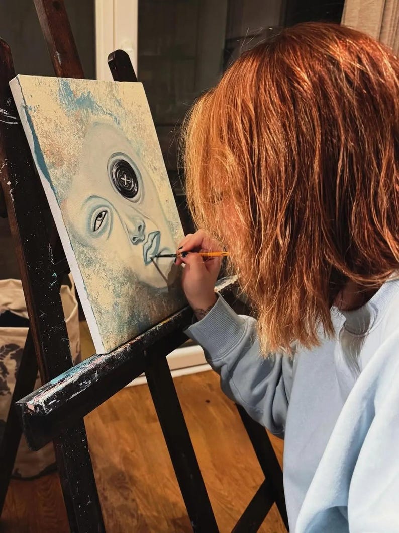 May include: A rectangular canvas painting on a black wooden easel features a stylized face with a prominent black button replacing one eye. The artwork uses shades of pale blue, cream, and white, with a fine brush adding detail to the lips.