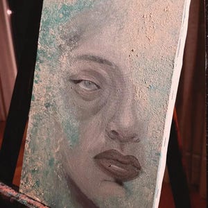 May include: A rectangular canvas painting featuring a close-up portrait of a face with dark lips and defined eyes. The artwork uses a textured application of paint in shades of teal, beige, and grey, creating a weathered, layered appearance.