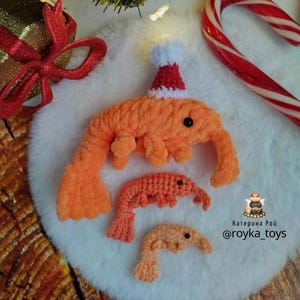 May include: Three crocheted orange shrimp ornaments, one wearing a red and white Santa hat. The ornaments are on a white fluffy surface. The text "Katerina Roy @royka_toys" is in the lower right corner of the image.