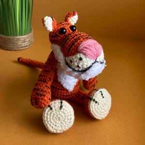 May include: A handmade orange and black crochet tiger doll with a pink nose, white muzzle, and white accents on the ears. The tiger has black eyes and is sitting with its legs extended. The doll is a cute, soft toy.
