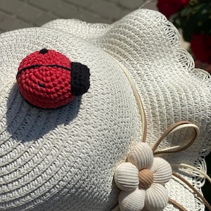 Puede incluir: Un sombrero de sol tejido en color blanco que presenta un pequeño adorno de mariquita de ganchillo en rojo y negro sobre la copa y una flor de tela blanca con centro beige sujeta al lateral con finos cordones decorativos marrón claro.