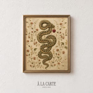 May include: A framed wall art print featuring a central green snake with orange and yellow spots, coiled in an S-shape, holding a small red apple in its mouth. The background is a cream-coloured fabric texture with delicate floral vines.