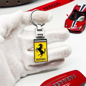 May include: A Ferrari keychain with a yellow and black emblem, held by a gloved hand. A red Ferrari car model and a red car key are also visible. A red ribbon with the Ferrari logo is in the background.