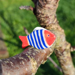 Peut inclure: Ornement en forme de poisson en bois, avec une tête et une queue rouges, et un corps rayé bleu et blanc. Le poisson est posé sur une branche d'arbre marron, avec de l'herbe verte en arrière-plan.