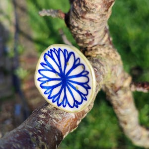 Peut inclure: Un petit pendentif rond en céramique avec un motif floral bleu. Le pendentif est blanc avec un motif de fleur bleue et un bord doré. Il repose sur une branche d'arbre marron, avec un arrière-plan vert flou.